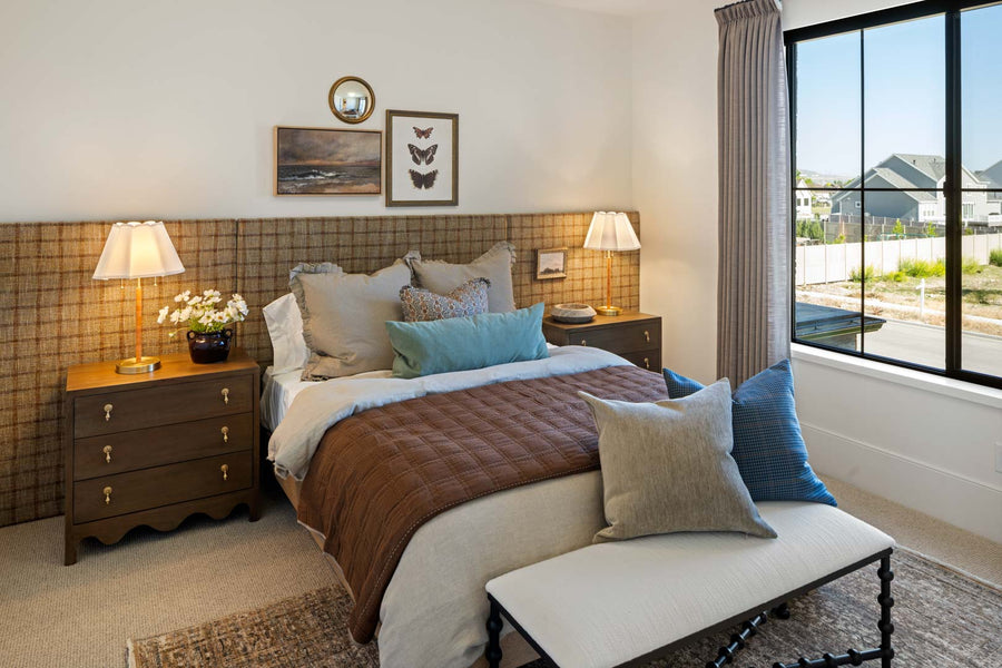 A styled bedroom interior featuring a bed with layered brown and blue bedding, matching nightstands with lamps, a plaid headboard, and a large window with a view of neighboring houses.