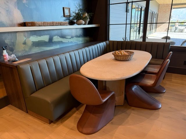 A modern dining nook featuring a curved upholstered booth in dark olive green, a round light beige stone-top table, and four sculptural brown chairs, set in a contemporary interior with wood floors and a large window.