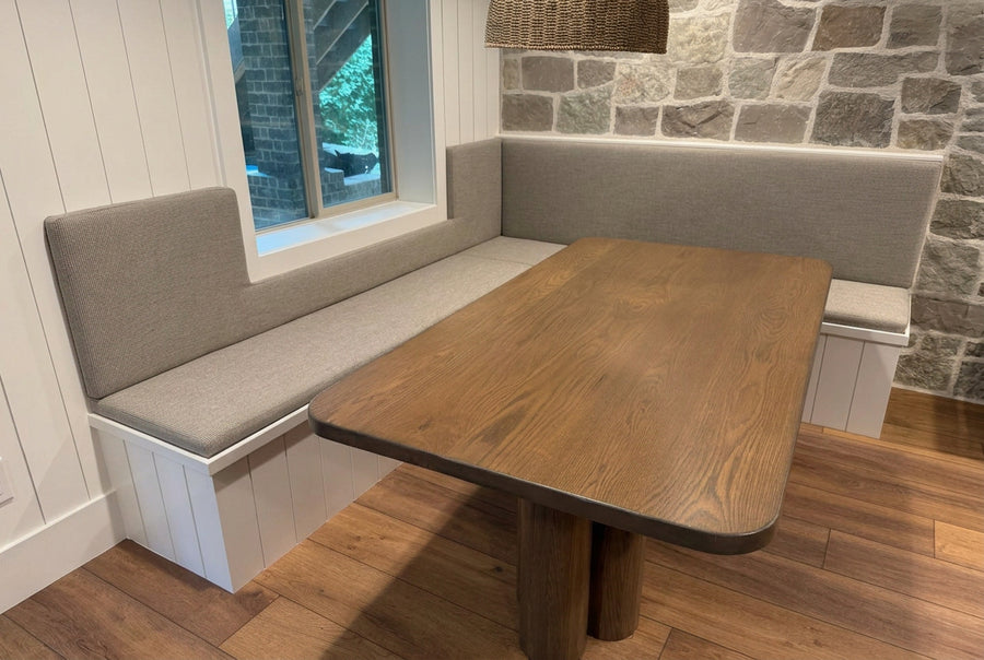 A wooden dining table with a colorful jigsaw puzzle on top, set in a modern L-shaped upholstered banquette seating area with a stone wall and wood floor.
