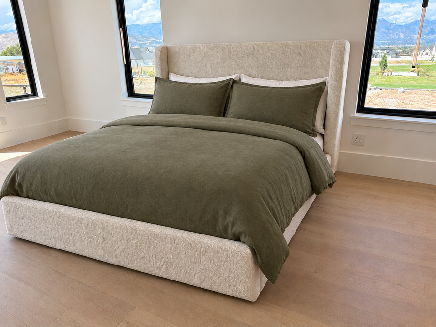An upholstered bed in a bright bedroom, dressed with an olive green duvet cover and two matching pillow shams. The bed has a tall, wingback-style upholstered headboard and base in a light beige fabric.