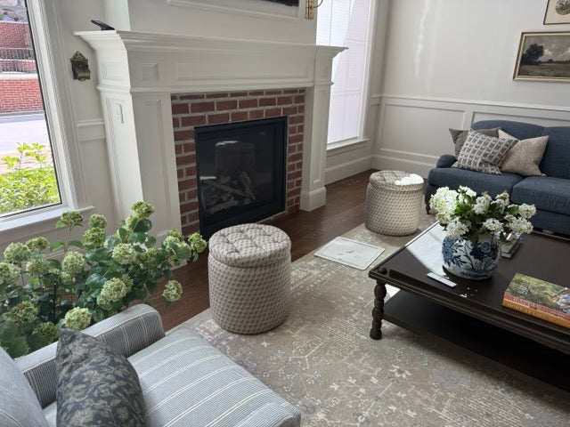 A cozy living room scene featuring a brick fireplace with a white mantel, a blue sofa with patterned pillows, two round woven ottomans, a dark wood coffee table with flowers, and a striped armchair in the foreground.