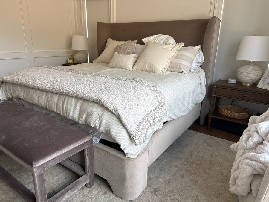 A styled bedroom scene featuring a bed with a tall upholstered headboard, cream bedding, multiple decorative pillows, a matching upholstered bed skirt, a wooden nightstand with a lamp, and a bench at the foot of the bed.