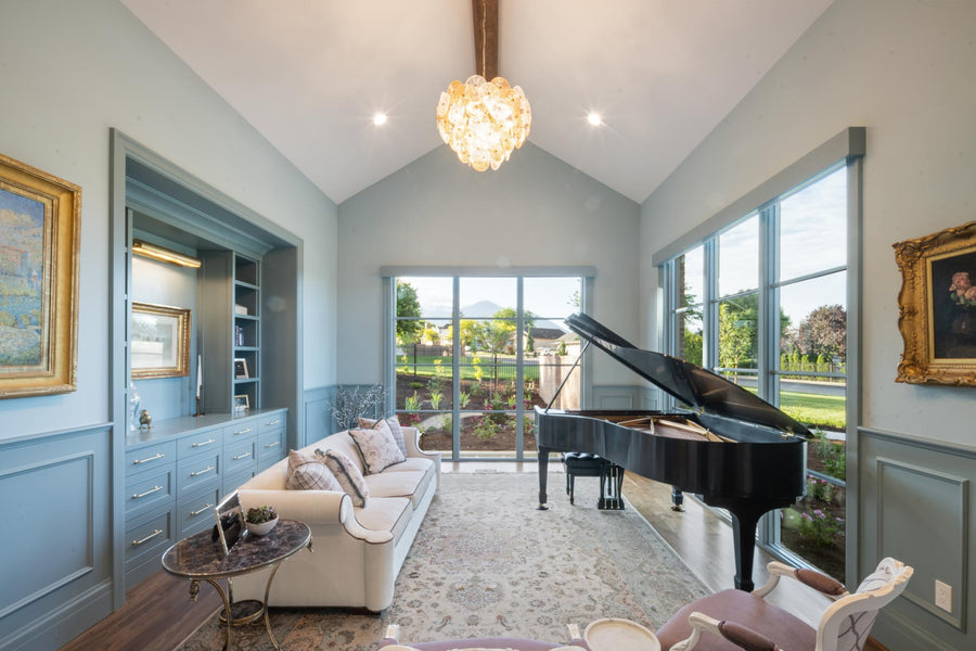A bright, elegant living room with light blue walls, featuring a black grand piano, a cream sofa, a patterned rug, and a glowing crystal chandelier. Large windows look out onto a landscaped garden.