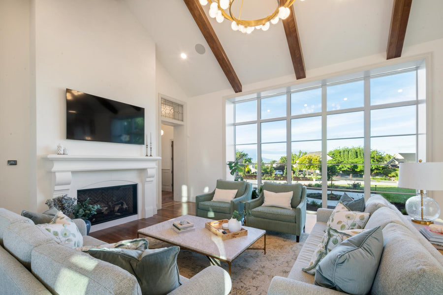 A bright, modern living room with high ceilings, exposed wood beams, a fireplace with a mounted TV, large floor-to-ceiling windows, and multiple upholstered sofas, armchairs, and a coffee table.