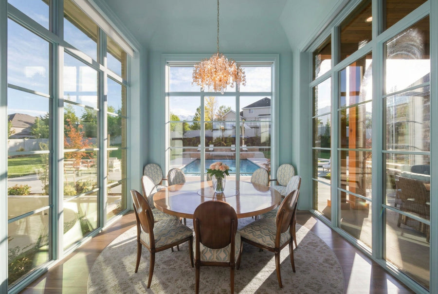 A bright sunroom dining area with a round wooden dining table, six upholstered wooden chairs, a hanging crystal chandelier, and large glass windows overlooking a pool and backyard.