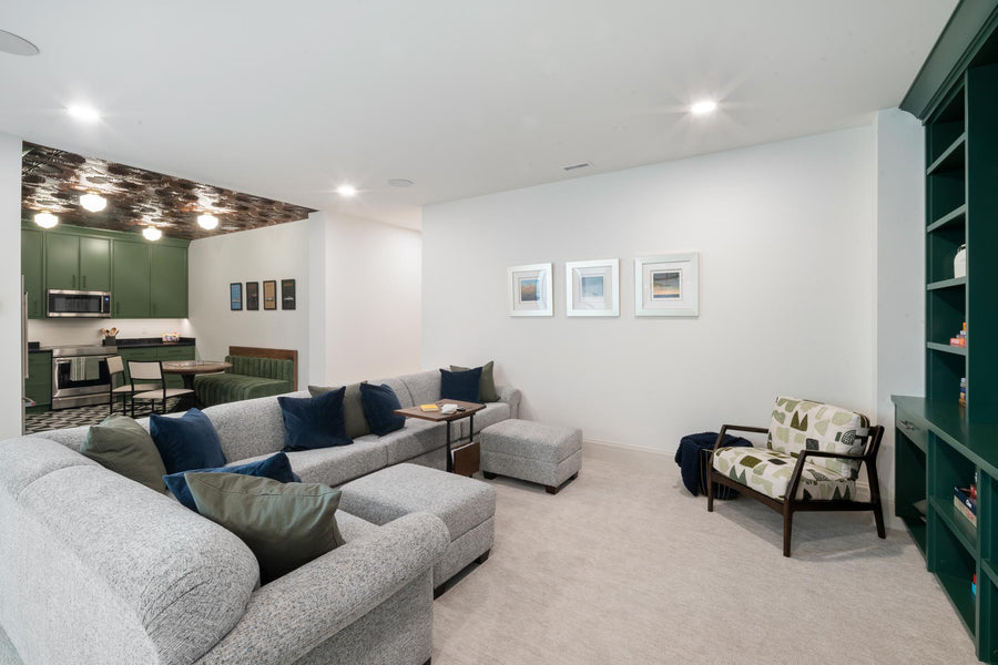 A modern living room featuring a large light gray sectional sofa with blue and olive accent pillows, a matching ottoman, a patterned armchair, and a dark green built-in shelving unit on the right.