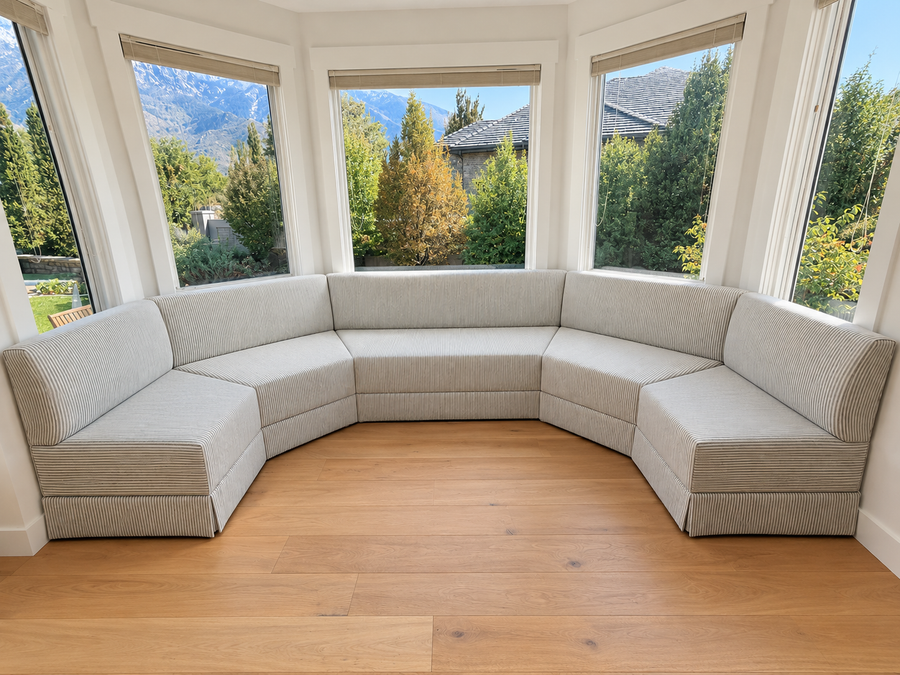 A curved, U-shaped upholstered corner sofa in a light beige ribbed fabric, set in a bright room with a wood floor and large windows looking out to trees and mountains.
