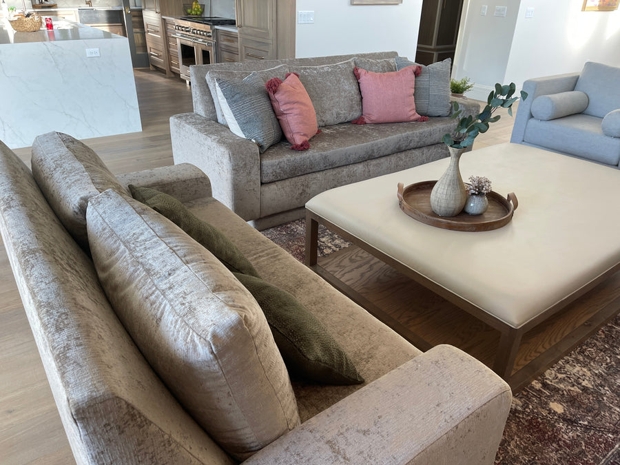 A large upholstered sofa in a textured light beige fabric. The sofa features thick, oversized back cushions and two pink throw pillows.