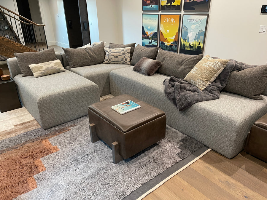 A modern L-shaped sectional sofa in textured light gray fabric, styled with multiple gray and beige throw pillows and a dark gray throw blanket. In front of the sofa is a brown leather ottoman.