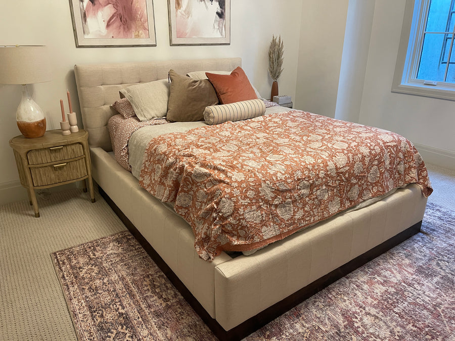 A bed with a tufted cream upholstered frame, dressed in a rust-orange and white patterned comforter, beige pillows, and a striped bolster, styled in a bedroom with a nightstand, lamp, and rug.