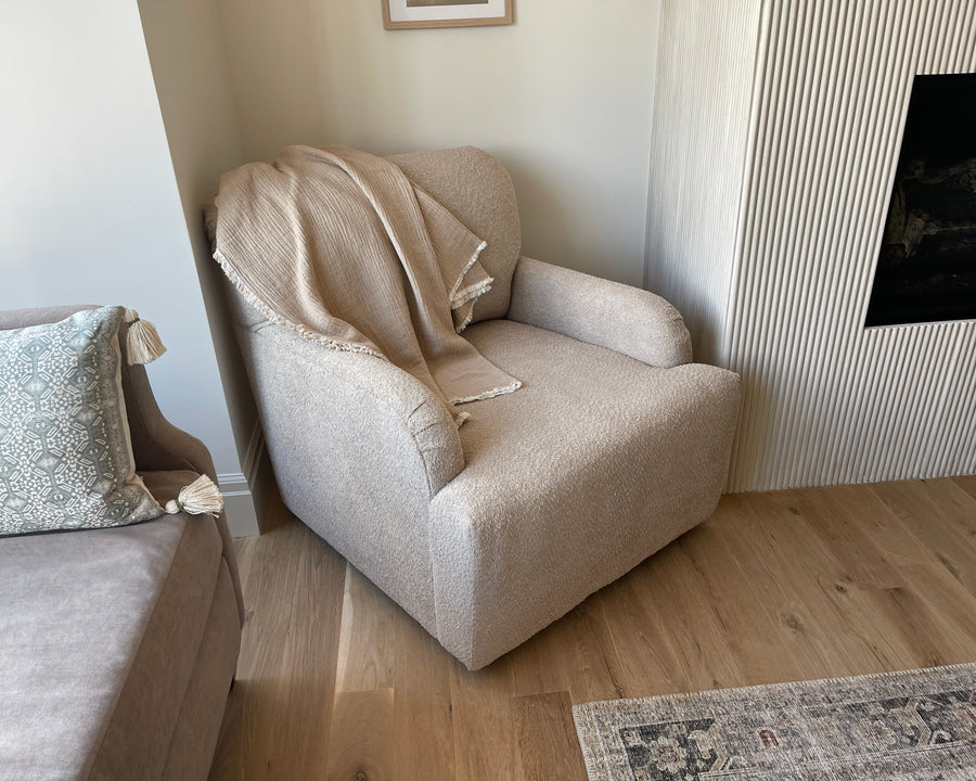 A beige upholstered armchair with a textured cream throw blanket draped over it, placed in a living room corner next to a sofa and a fluted wall panel with a fireplace.
