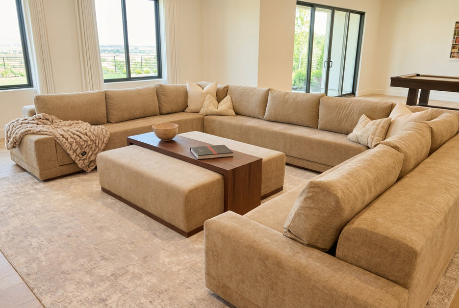A modern sectional sofa in a light beige fabric in a living room setting with a matching coffee table ottoman and textured rug.