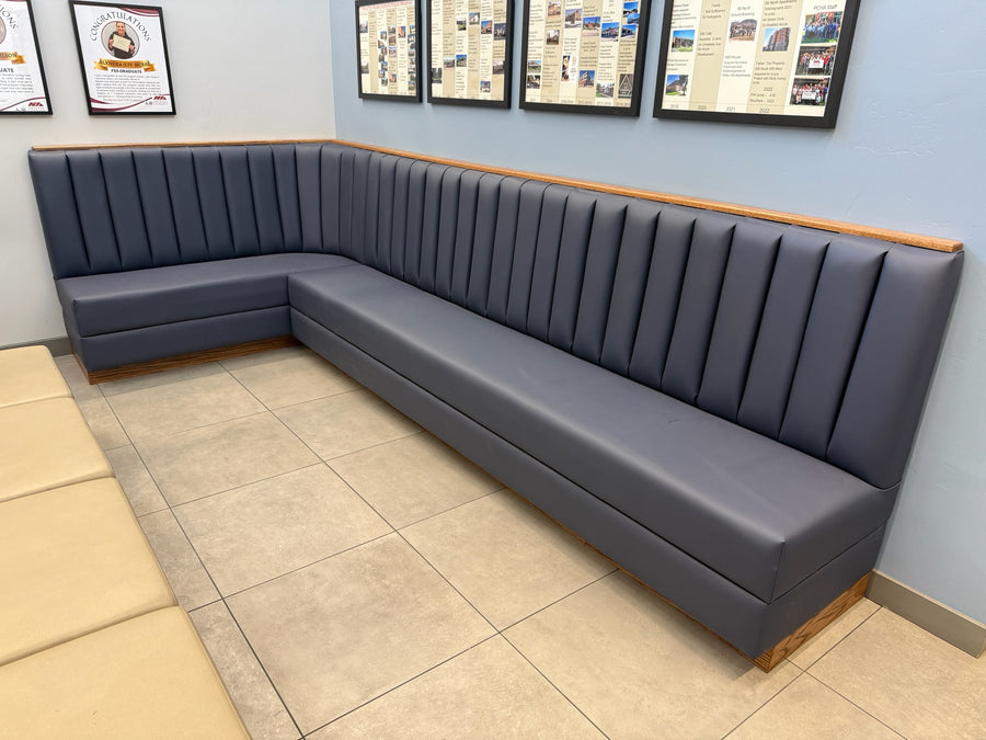 A dark gray upholstered L-shaped booth with vertical channel tufting and light wood trim, installed in a restaurant or cafe setting with tiled floors and framed wall displays.