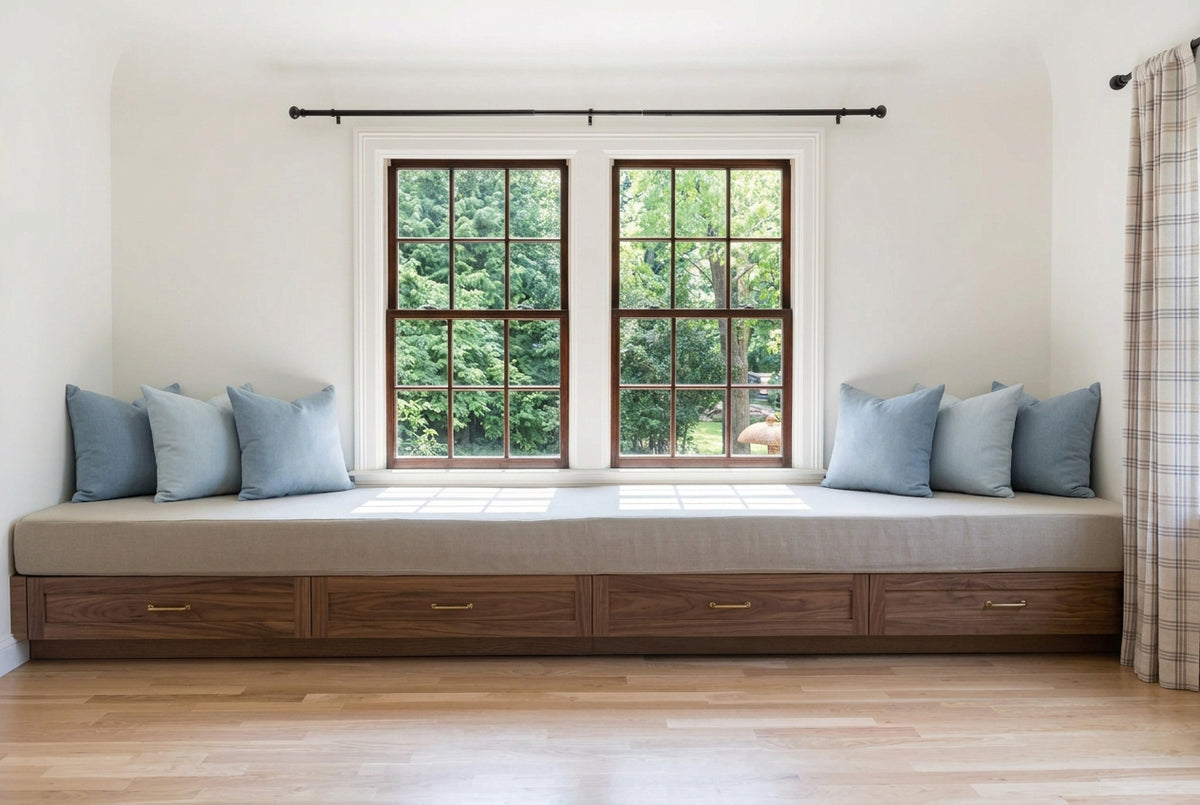 A long upholstered window seat cushion with a wooden base and several matching pillows, installed along one wall in a bright room with wood floors and large multi-pane windows.