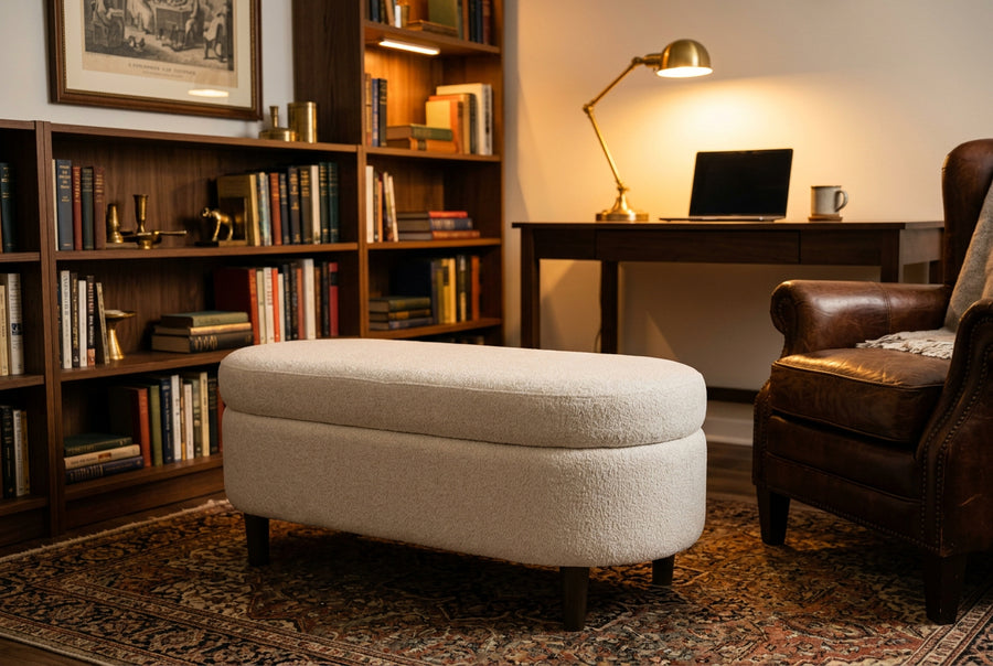 A cream-colored bouclé ottoman with dark wooden legs, styled in a cozy home office setting with a dark wood desk, leather armchair, and bookshelves in the background.