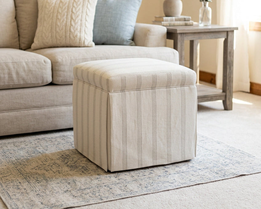 A square upholstered ottoman with a striped pattern in off-white and light gray, shown on a carpeted floor against a plain wall.