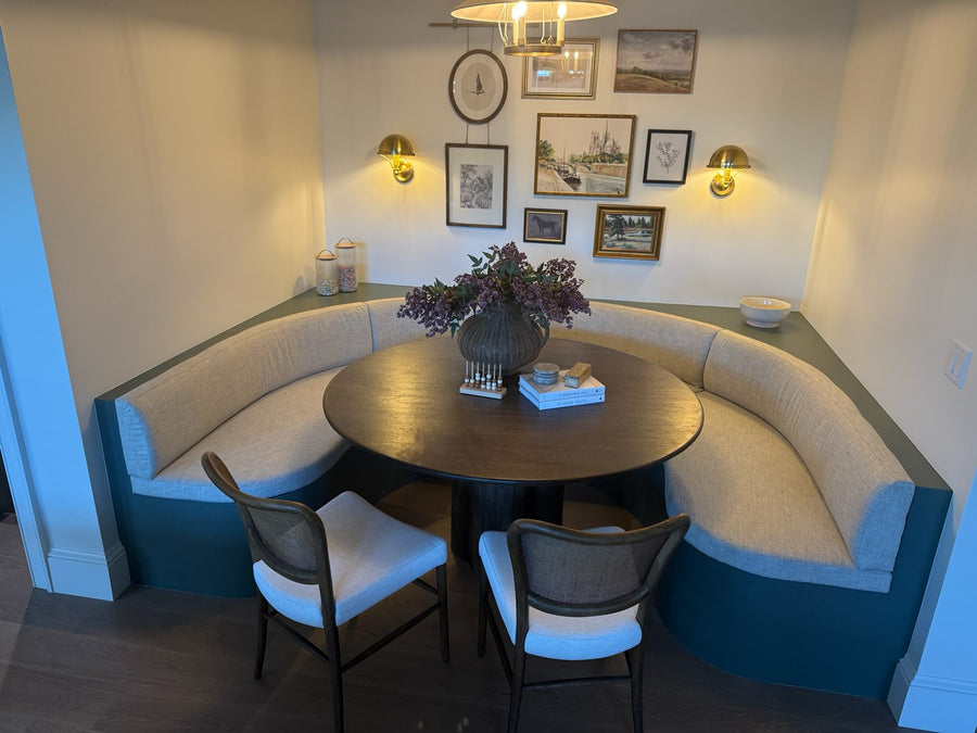 A cozy corner dining nook with a curved upholstered bench, a round dark wood table, two chairs with white seat cushions, and a floral centerpiece, set against a wall with framed artwork and wall sconces.