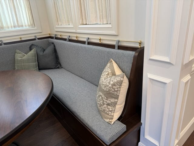 A corner upholstered banquette seating bench with dark wood framing, gray textured fabric cushions, and decorative throw pillows in a bright interior setting.